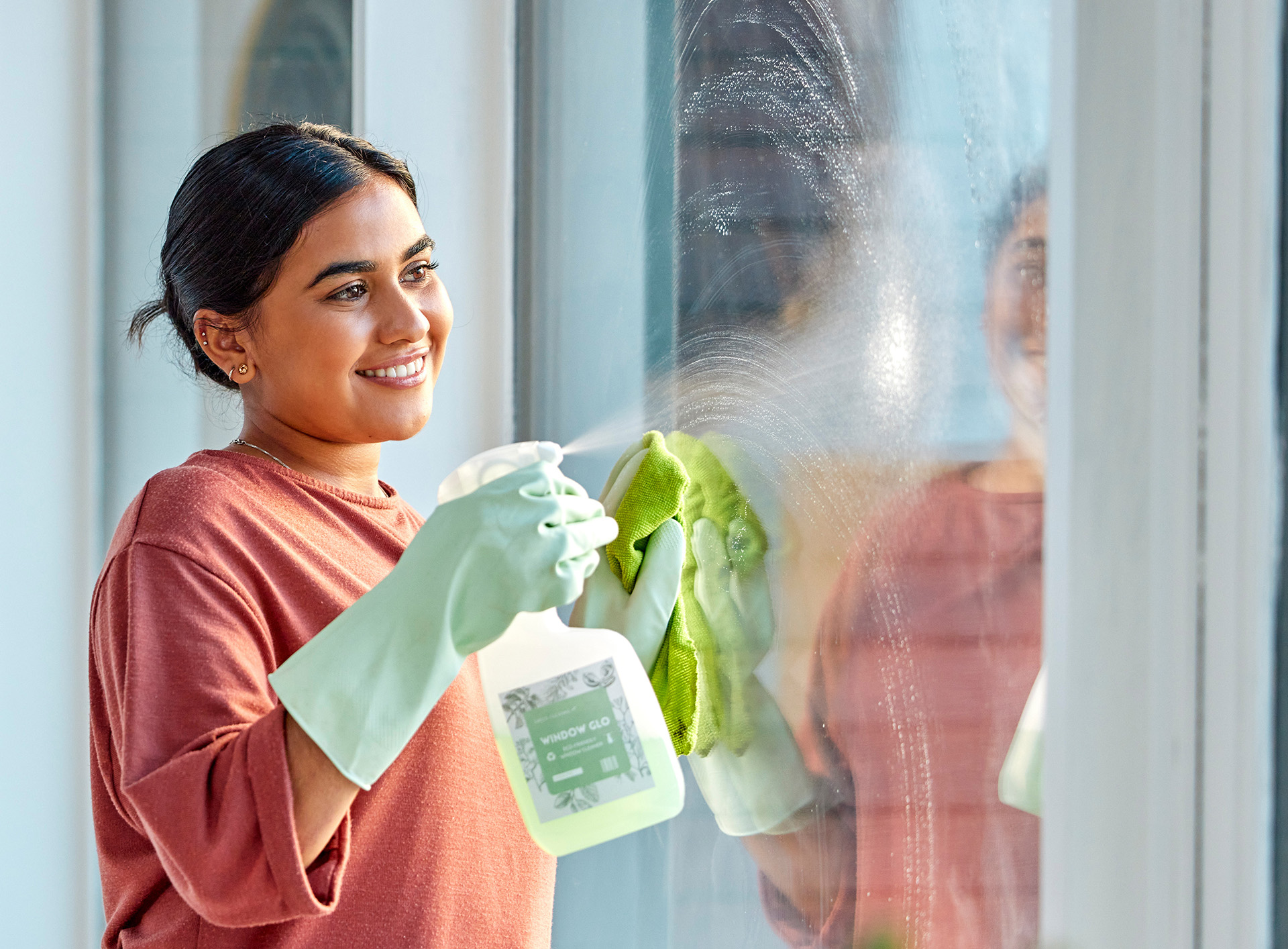 Smiling person wearing gloves cleans windows with spray and cloth. The window reflects their image, creating a cheerful and tidy atmosphere.