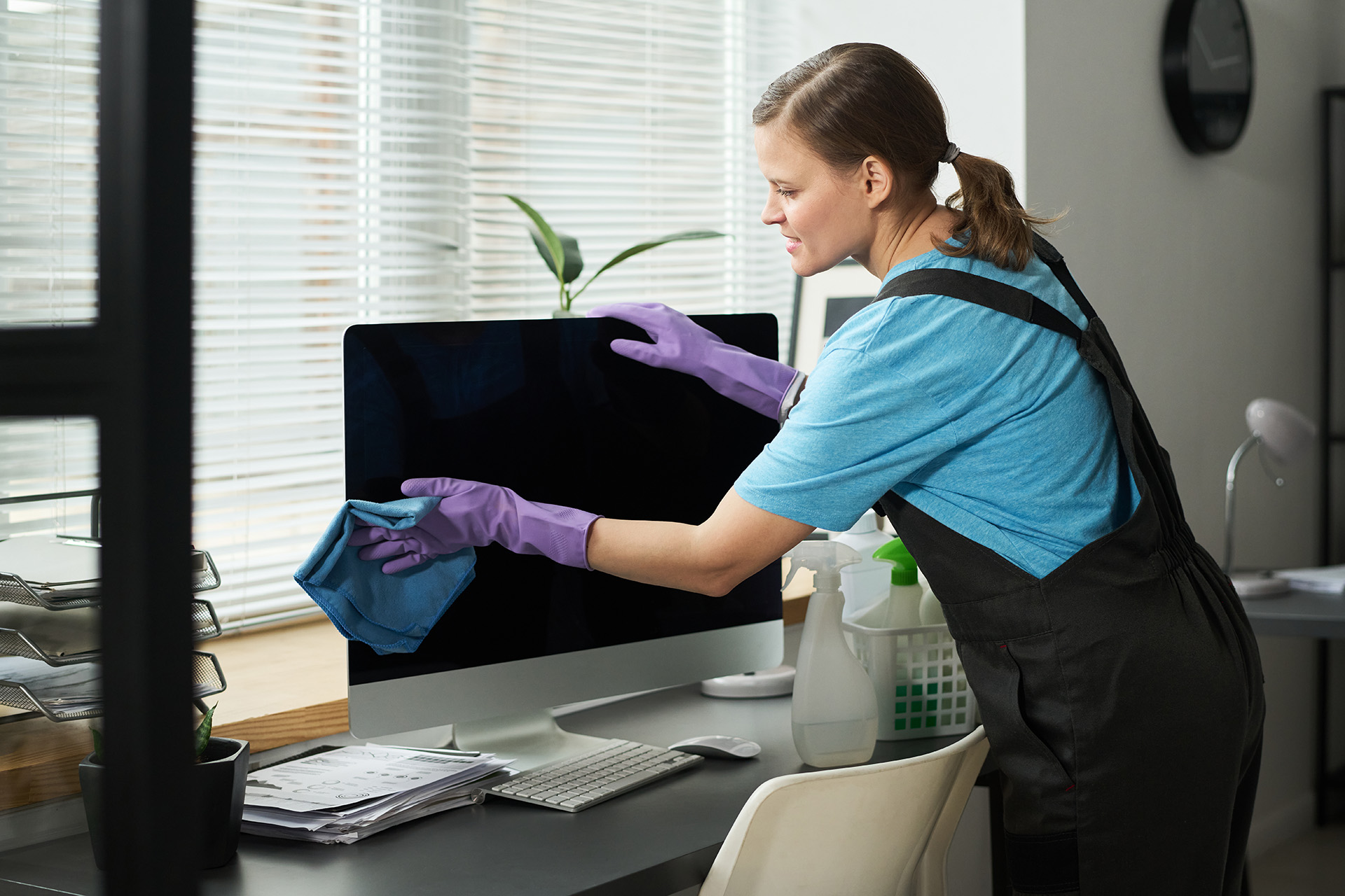 A person wearing gloves uses a cloth to clean a computer monitor in an office setting. Cleaning supplies and paperwork are visible on the desk.