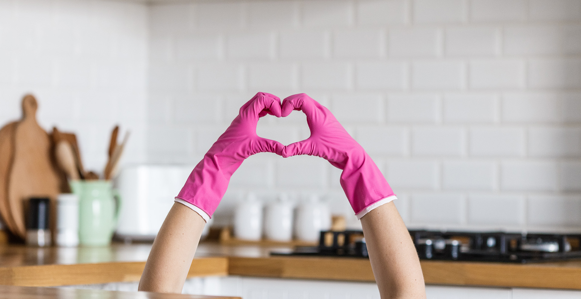 Hands in pink rubber gloves form a heart shape against a blurred kitchen background with white tiles, conveying a playful, caring tone.